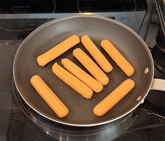 While the water heats, warm a frying pan over medium heat and coat it lightly with olive oil spray. Add the hot dogs and cook 6 to 7 minutes, turning frequently, until every side is lightly browned. Cut them into bite sized pieces and set aside.