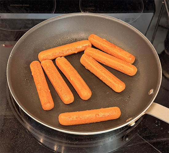 While the water heats, warm a frying pan over medium heat and coat it lightly with olive oil spray. Add the hot dogs and cook 6 to 7 minutes, turning frequently, until every side is lightly browned. Cut them into bite sized pieces and set aside.